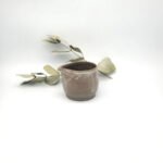 Handcrafted dark clay mini cream pitcher with a spouted rim resting beside eucalyptus leaves.
