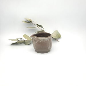 Handcrafted dark clay mini cream pitcher with a spouted rim resting beside eucalyptus leaves.